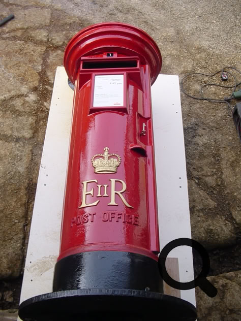 Roter Briefkasten mit goldener Krone und den Buchstaben "E II R", auf einem weißen Brett liegend.