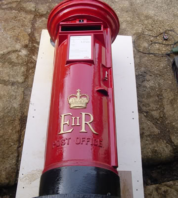 Roter, zylindrischer Briefkasten mit goldener Krone und den Buchstaben "E II R" darauf, liegend auf einer weißen Unterlage.