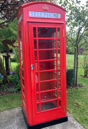 Rote Telefonzelle auf einem Gehweg, umgeben von Gras und Bäumen.