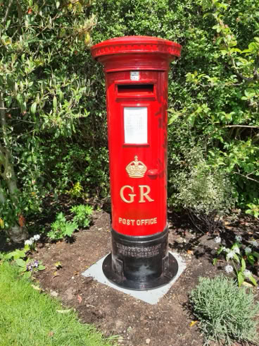 Roter Briefkasten mit der Aufschrift "GR" und Krone, umgeben von Pflanzen und Gras.