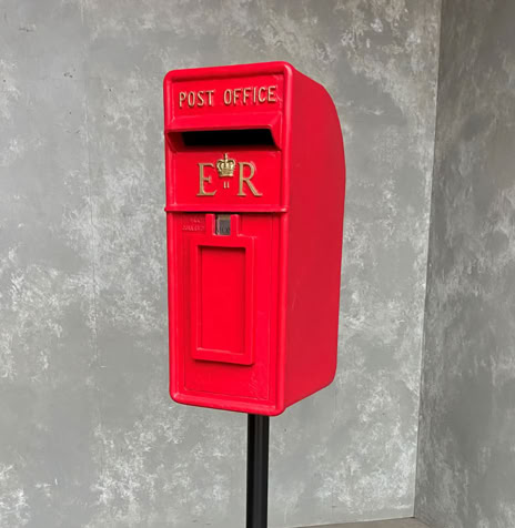 Roter Briefkasten auf einem schwarzen Ständer vor einer grauen Wand.